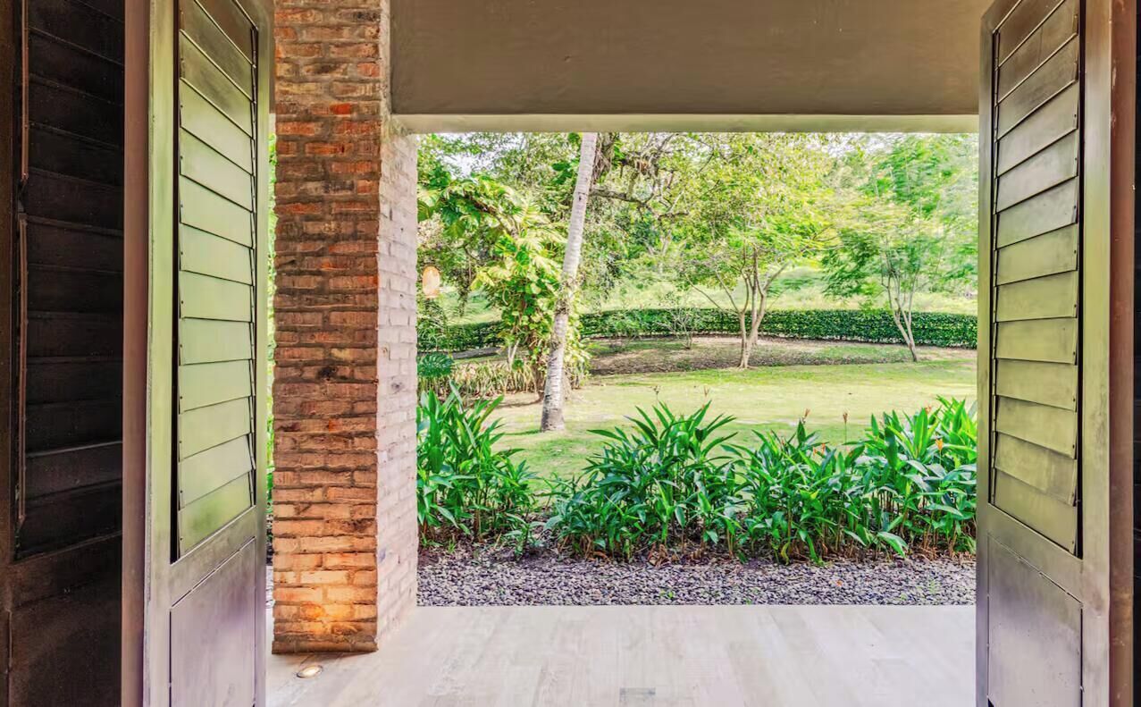 A covered outdoor area with a brick column, a paved floor, and a view of a landscaped garden with trees and shrubs.