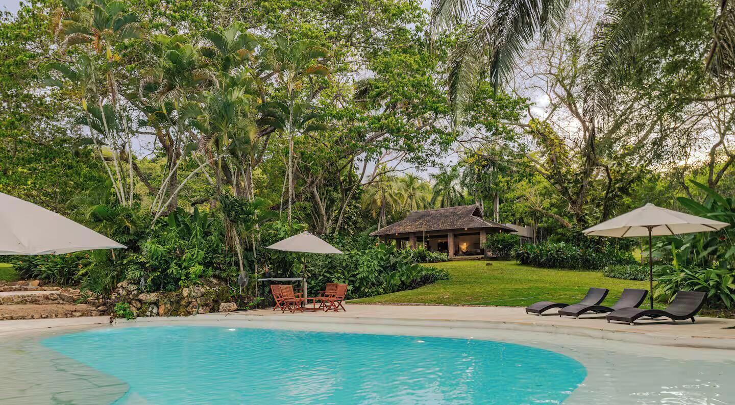 A swimming pool with lounge chairs and umbrellas, surrounded by lush greenery and a small building.
