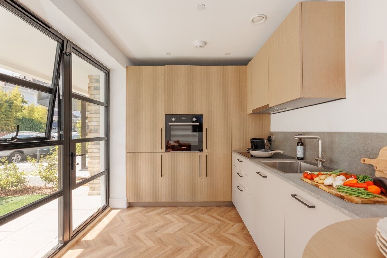 Une cuisine moderne équipée d’un four encastré, d’un évier et d’une fenêtre donnant sur un quartier résidentiel.