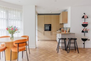 A modern kitchen with wooden cabinets, a built-in oven, and a dining area with a round table and orange chairs. - Sea Breeze by Grandeur Property (Poole)