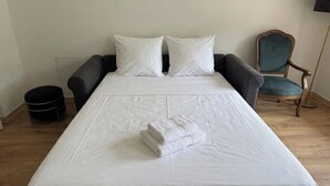 Ein ordentlich bezogenes Bett mit weißen Leinentüchern und einem gefalteten Handtuch, ein dunkler Sessel, ein Holzstuhl und ein Mülleimer in einem Hotelzimmer.
