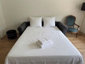 Ein ordentlich bezogenes Bett mit weißen Leinentüchern und einem gefalteten Handtuch, ein dunkler Sessel, ein Holzstuhl und ein Mülleimer in einem Hotelzimmer.