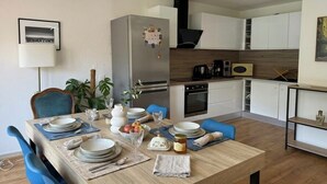Eine moderne Küche mit weißen Schränken, einem Edelstahlkühlschrank und einer Essecke mit blauen Stühlen und einem Holztisch, gedeckt für vier Personen.