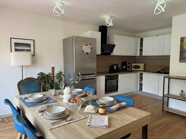 Una cocina moderna con armarios blancos, un refrigerador de acero inoxidable y un comedor con sillas azules y una mesa de madera preparada para cuatro.