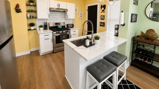 A kitchen with white cabinets, a stainless steel refrigerator, a stove, and a sink with a modern faucet. There are two bar stools and a wine rack with bottles.