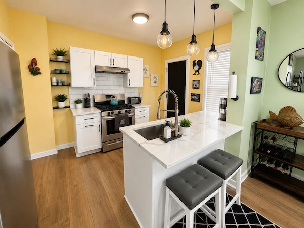 A kitchen with white cabinets, a stainless steel refrigerator, a stove, and a sink with a modern faucet. There are two bar stools and a wine rack with bottles.