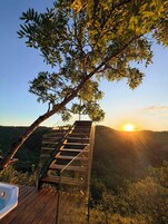 Um deck de madeira com guarda-corpo de vidro, com vista para uma paisagem arborizada ao pôr do sol.