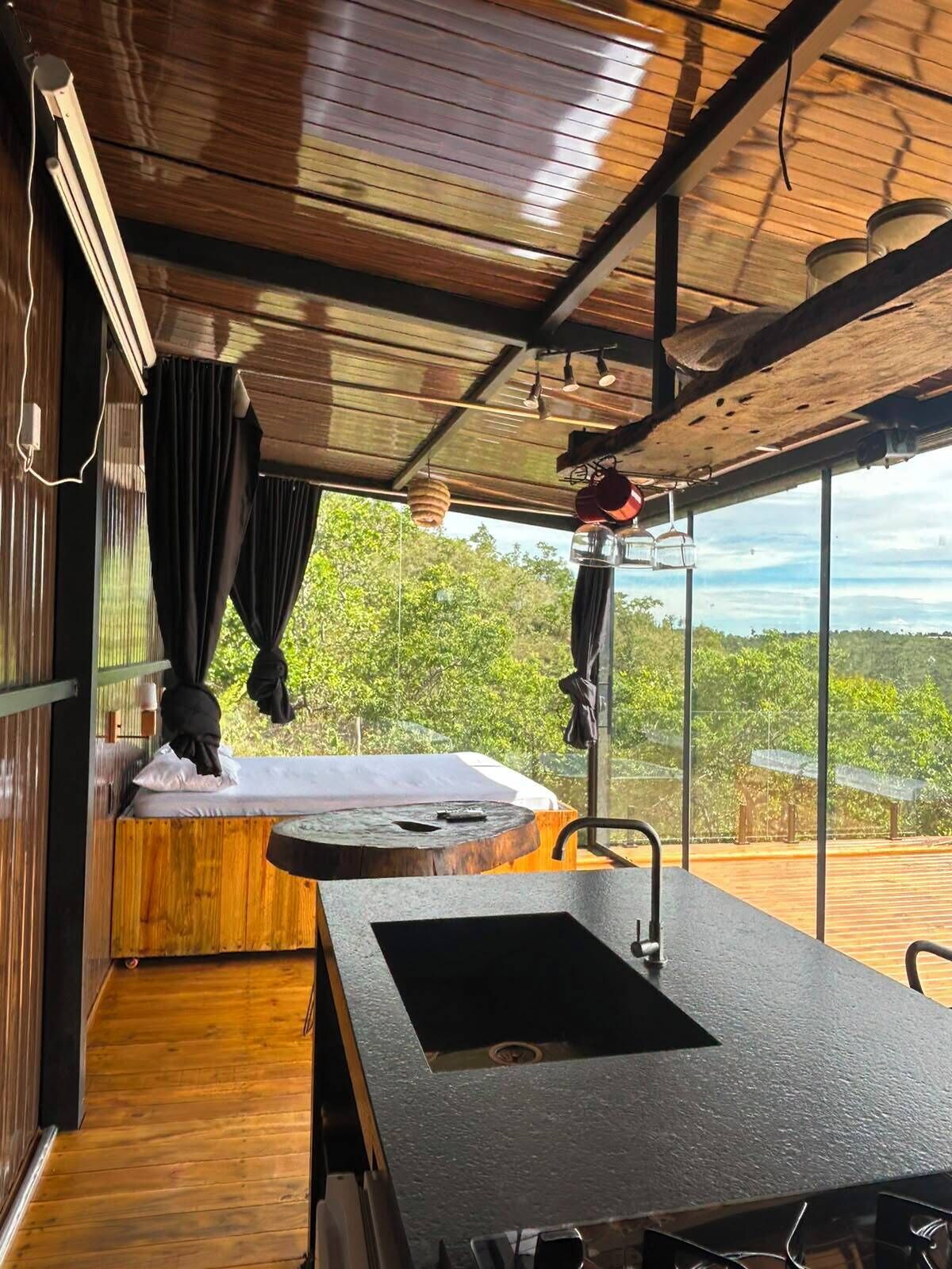 Habitación con piso de madera, un ventanal grande, cama, lavamanos y vistas al exterior.