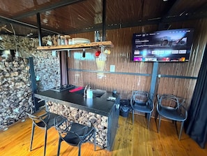 Zona de bar con pared de piedra, techo de madera y una televisión de pantalla plana mostrando un noticiero.