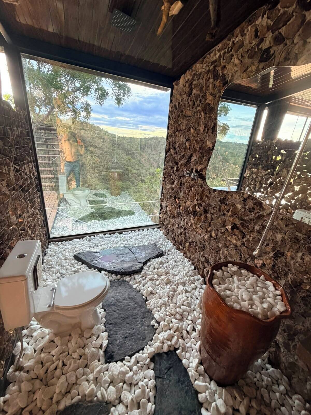 A bathroom with a stone wall, a large window, a toilet, and a stone basin.