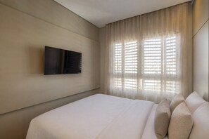 Ein Hotelzimmer mit Bett, Flachbildfernseher an der Wand und Fenster mit durchsichtigen Vorhängen.