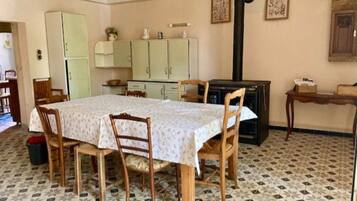 Una zona de menjador amb una taula preparada per a quatre persones, envoltada de caires de fusta. Hi ha una cuina al fons amb armaris i una estufa.