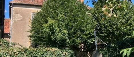 Un edificio con tejado de tejas rojas, rodeado de abundante vegetación y un muro de piedra.