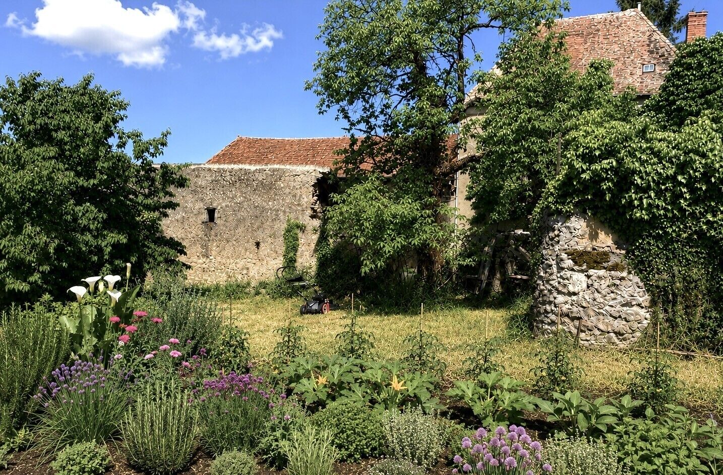 A stone building with a tiled roof, surrounded by lush greenery and flowering plants.