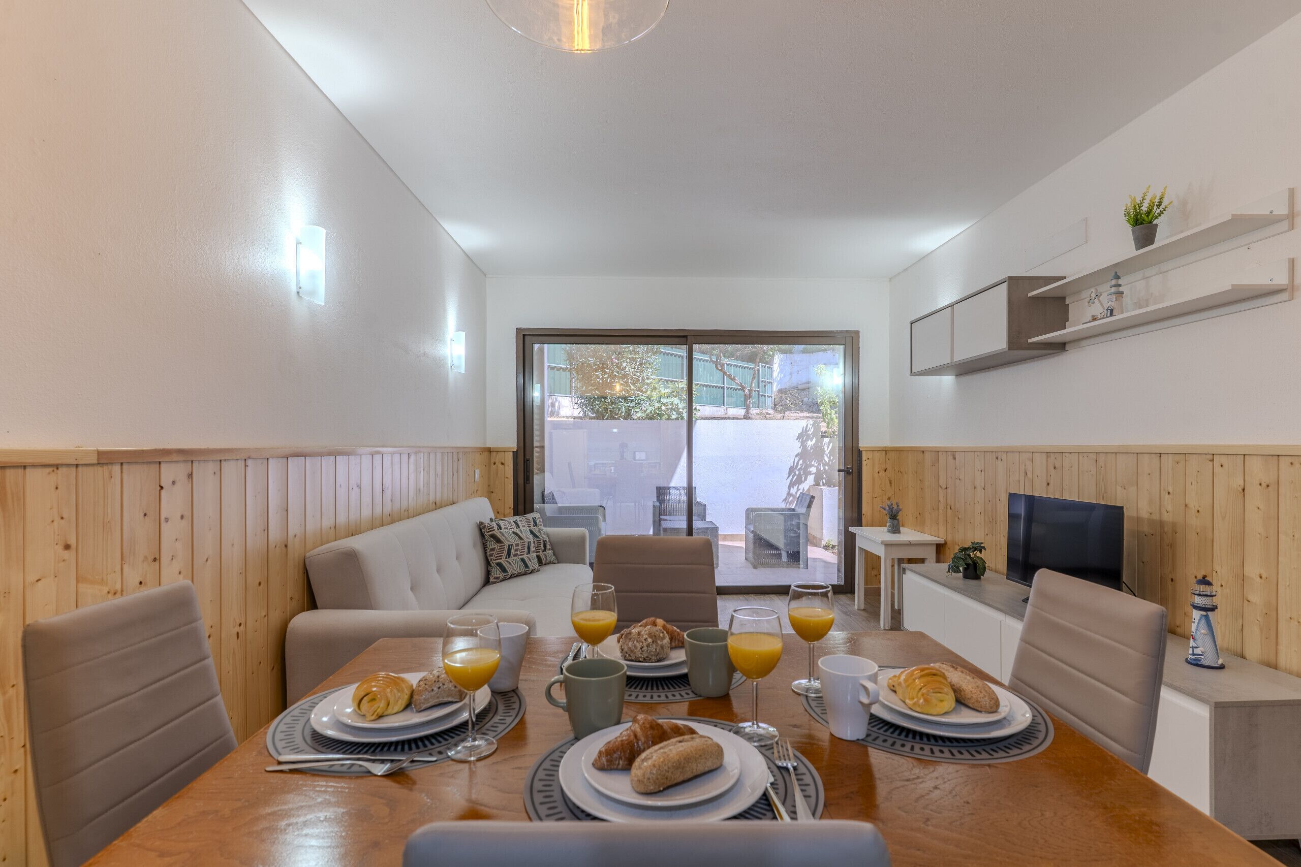 A dining area with a wooden table set for breakfast, a sofa, and a TV on a stand.