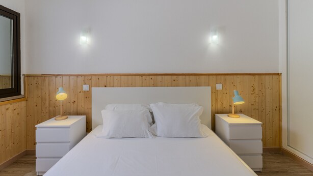 A bedroom with a wooden headboard, two bedside tables with lamps, a bed with white bedding, and a mirror on the left wall. - #400 Central Albufeira Oasis | 1BR & Pool (Albufeira)