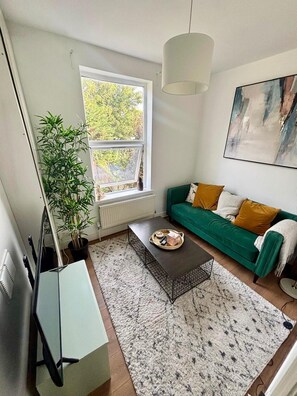A living room with a green sofa, a coffee table, a potted plant, and a window with a view of trees. - Apartment Sleeps 4 With Parking (London)