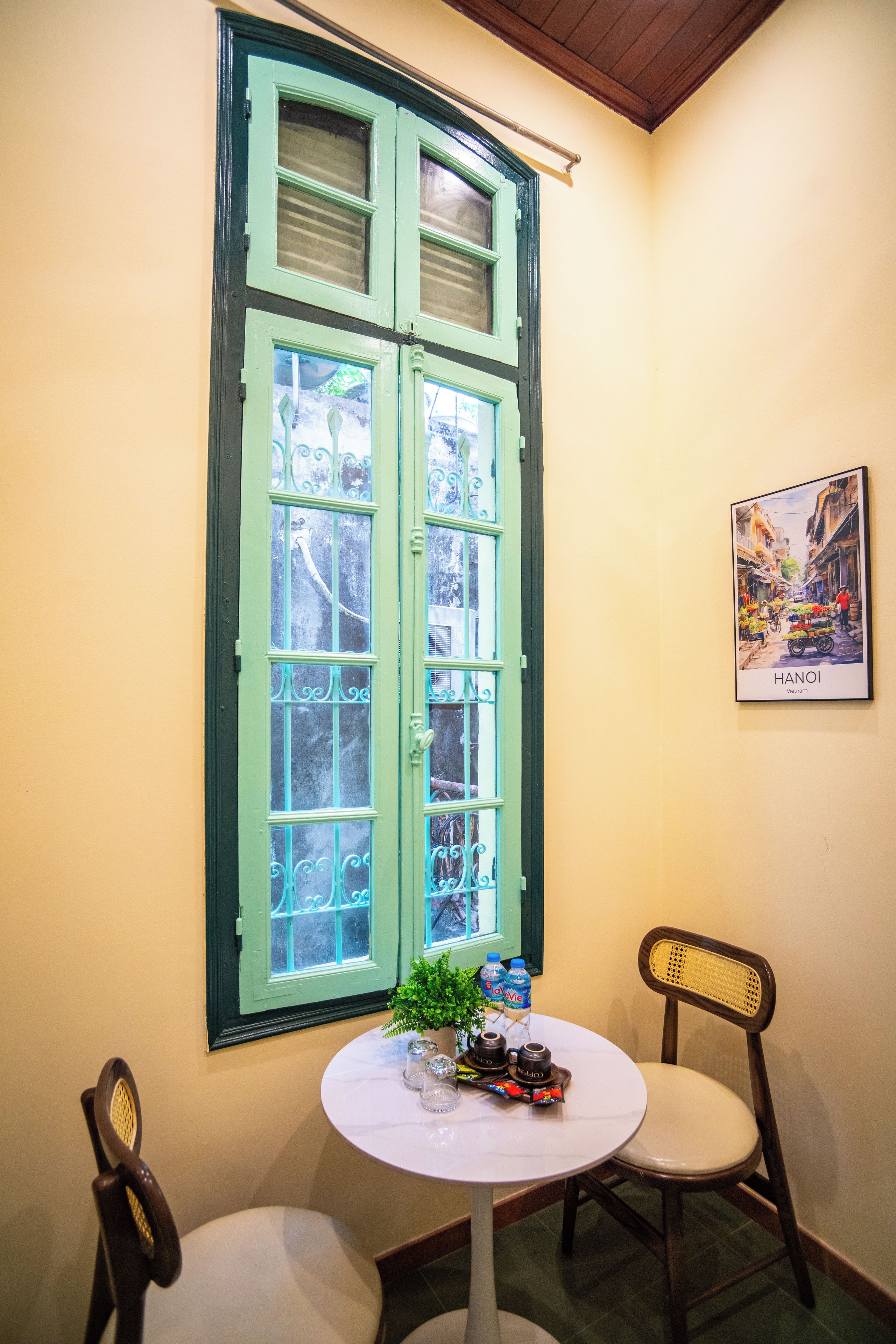 Ein Zimmer mit einem großen Fenster, einem kleinen runden Tisch mit Pflanze und Wasserflaschen, zwei Stühlen und einem gerahmten Bild an der Wand.