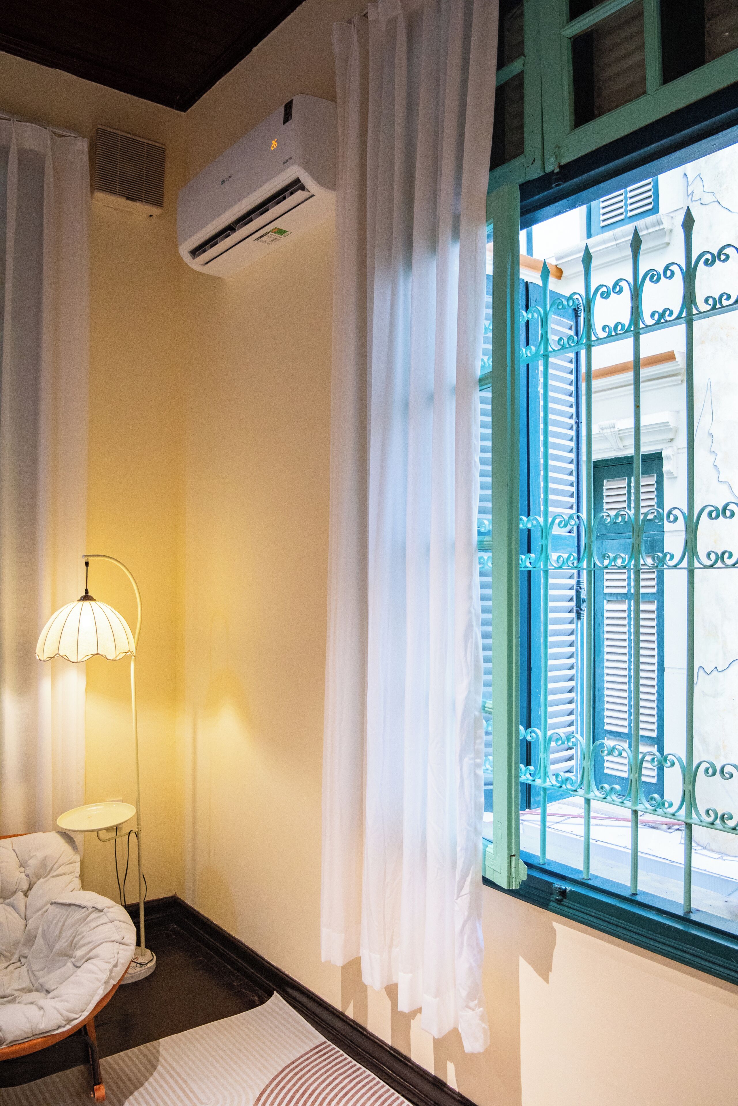 Una habitación con cama, mesita de noche, lámpara, ventana con rejas decorativas y equipo de aire acondicionado.
