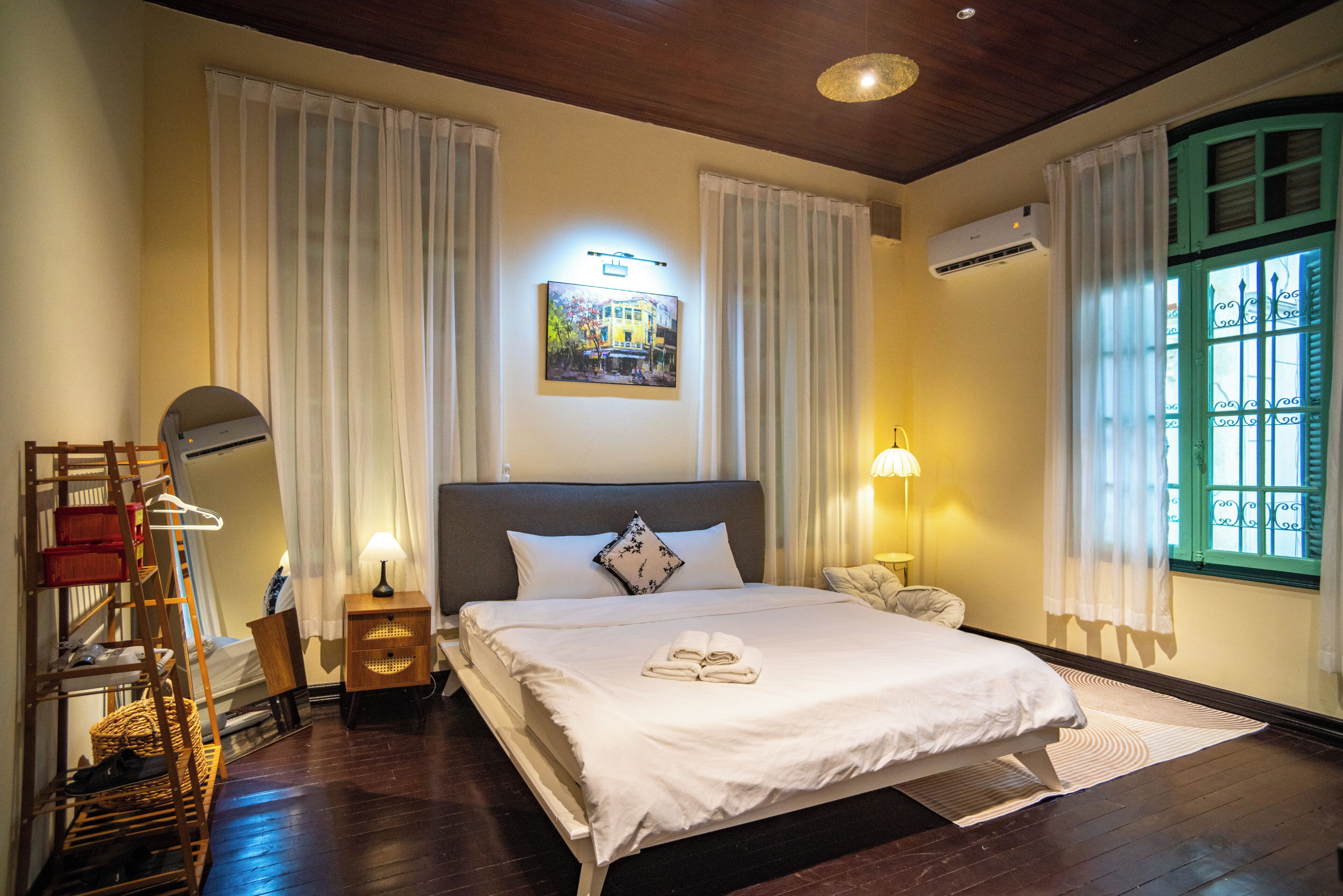 Quarto de hotel com uma cama grande, uma mesinha de cabeceira, um abajur, ar condicionado fixado na parede e uma janela com grades de ferro decorativas.