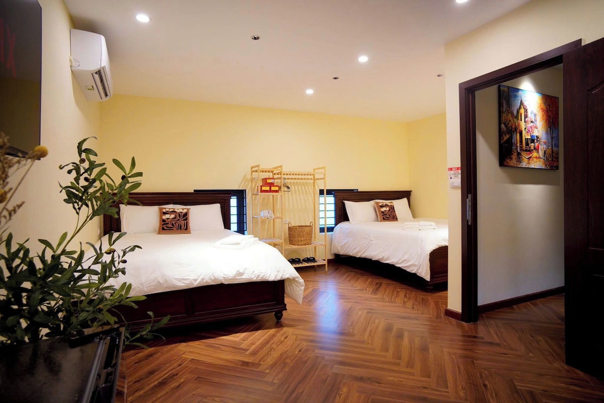 A hotel room with two beds, a wooden floor, a wall-mounted air conditioner, and a painting on the wall.