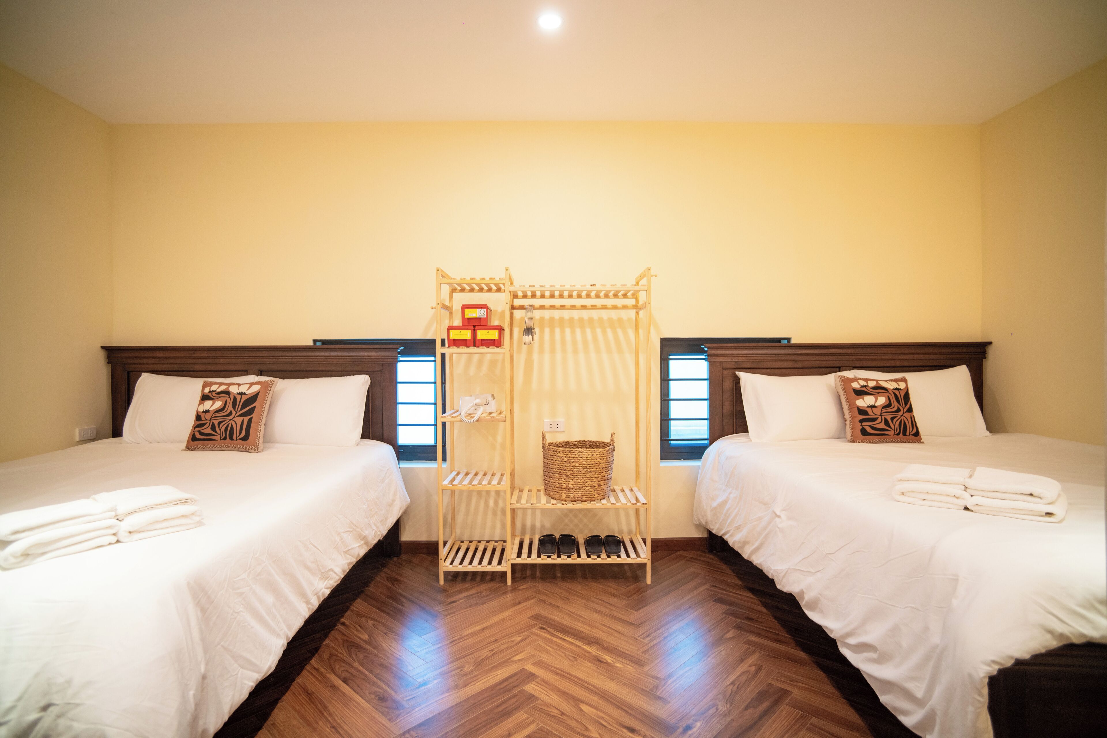 A hotel room with two beds, a wooden bedside table, a small shelf, and a window.