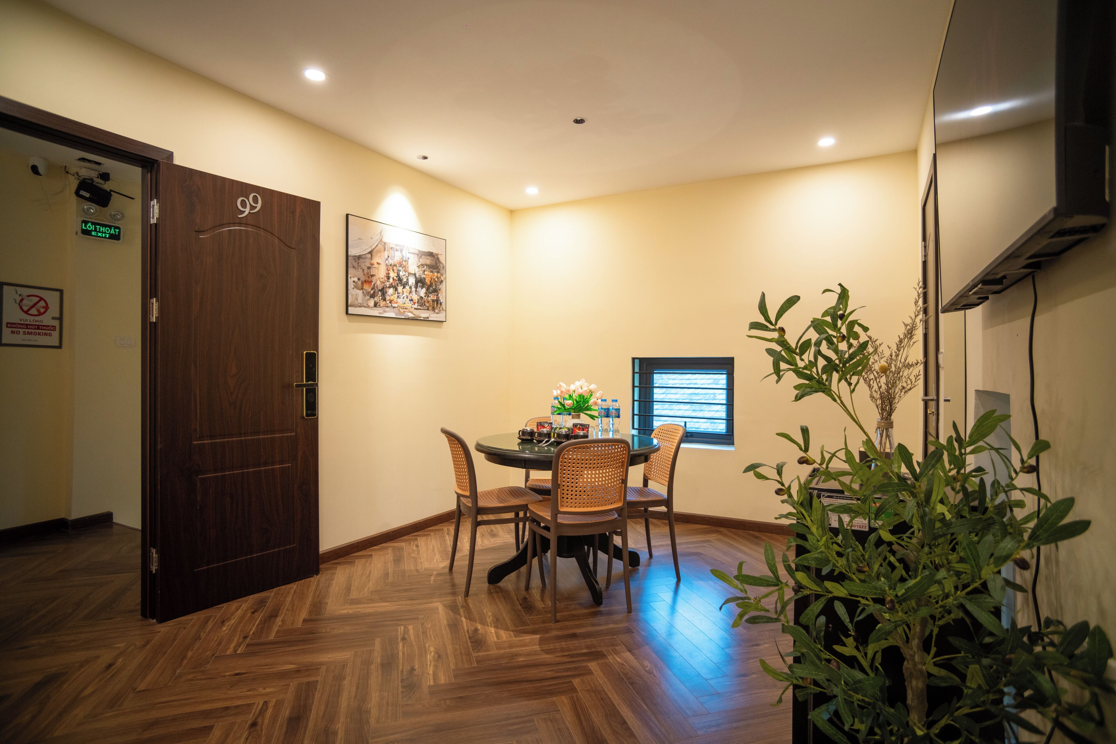 A hotel room with a wooden floor, a dining table with chairs, a painting on the wall, and a potted plant.