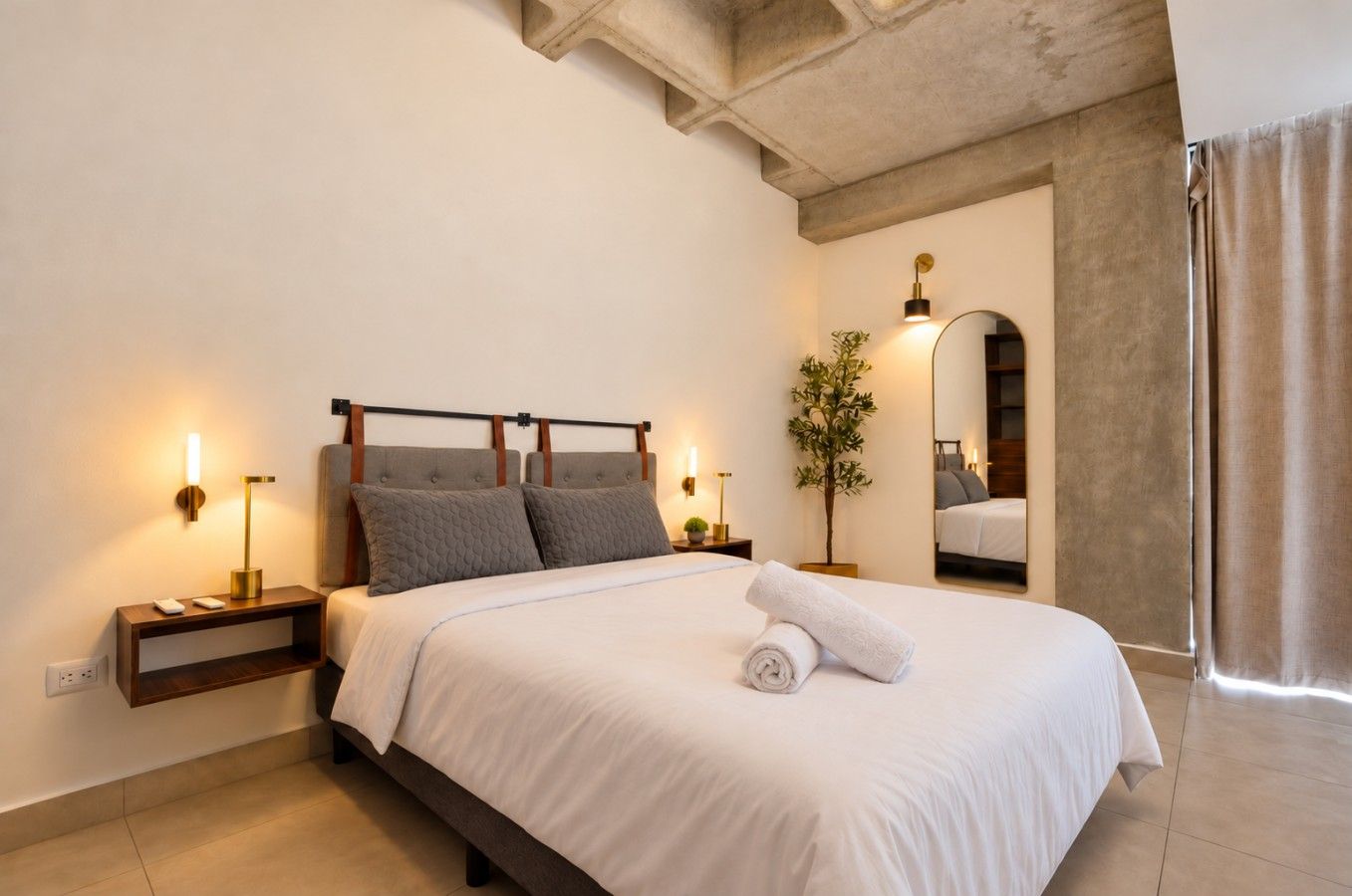 A hotel room with a bed, bedside lamps, a nightstand, a potted plant, and a mirror.