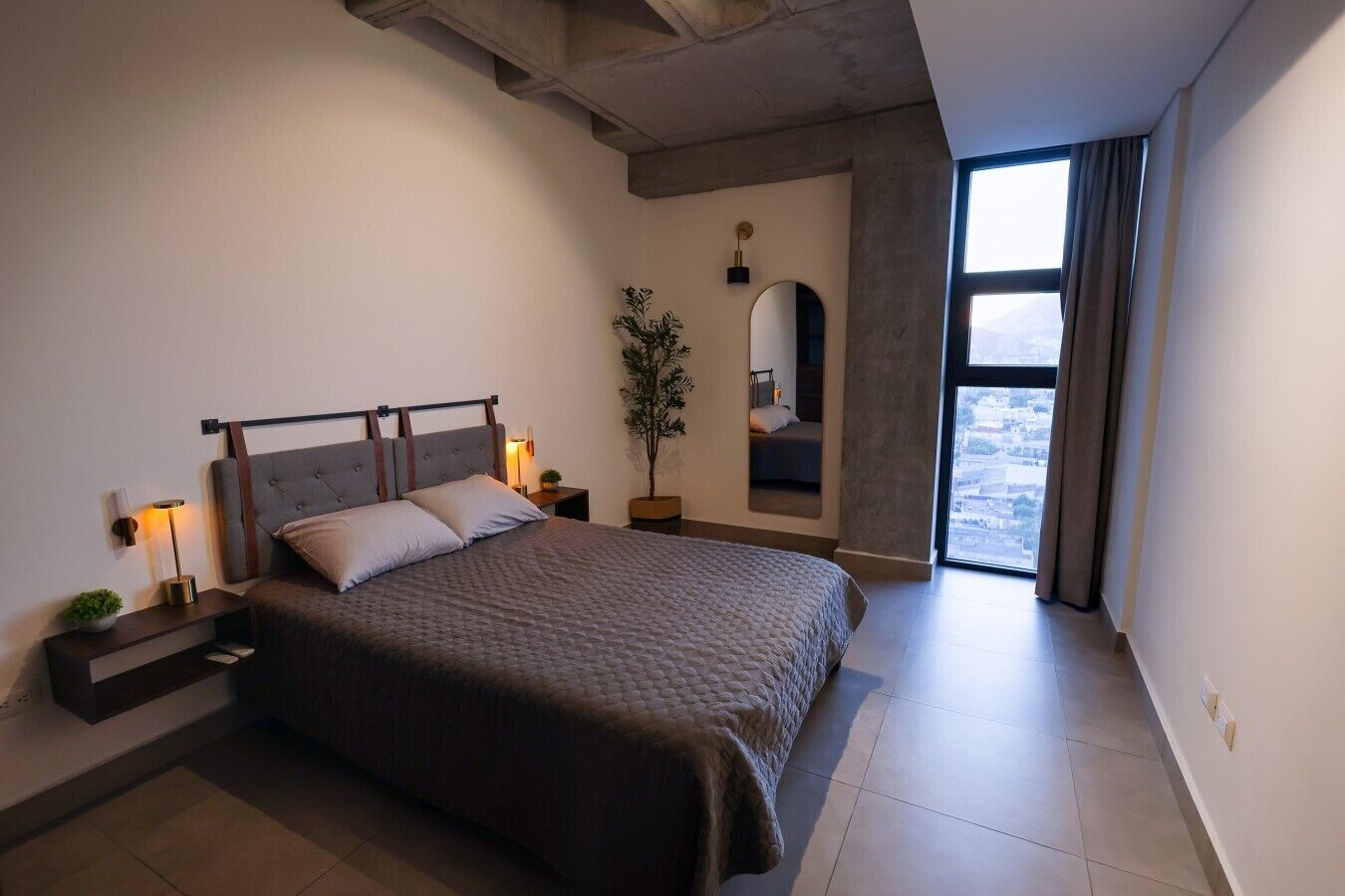 Une chambre moderne avec un grand lit, des lampes de chevet, une plante en pot et une vue par la fenêtre.