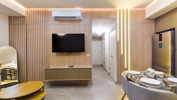 A modern hotel room with a flat-screen TV, a built-in air conditioner, a dining area with a glass table, and a minibar.