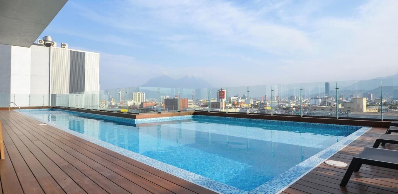 A rooftop swimming pool with a cityscape view, wooden deck, and glass railing.