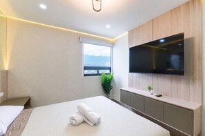 A modern hotel room with a large bed, a flat-screen TV mounted on the wall, a small window with a view, and a plant on the windowsill. - ¡Céntrico & Nuevo! Gym, Parking & Pool - 6G 2BR2br (Monterrey)