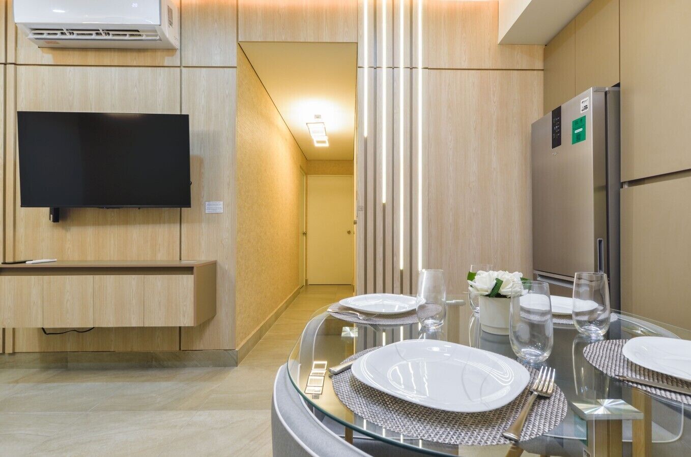 A modern hotel room with a flat-screen TV, a built-in refrigerator, and a dining area with a glass table set for two.