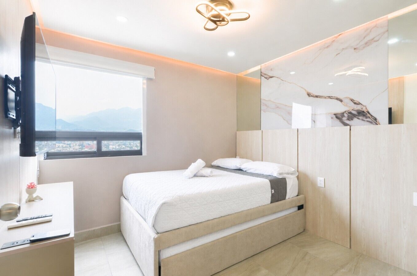 A modern bedroom with a bed, a desk, a TV, and a window with a city view.