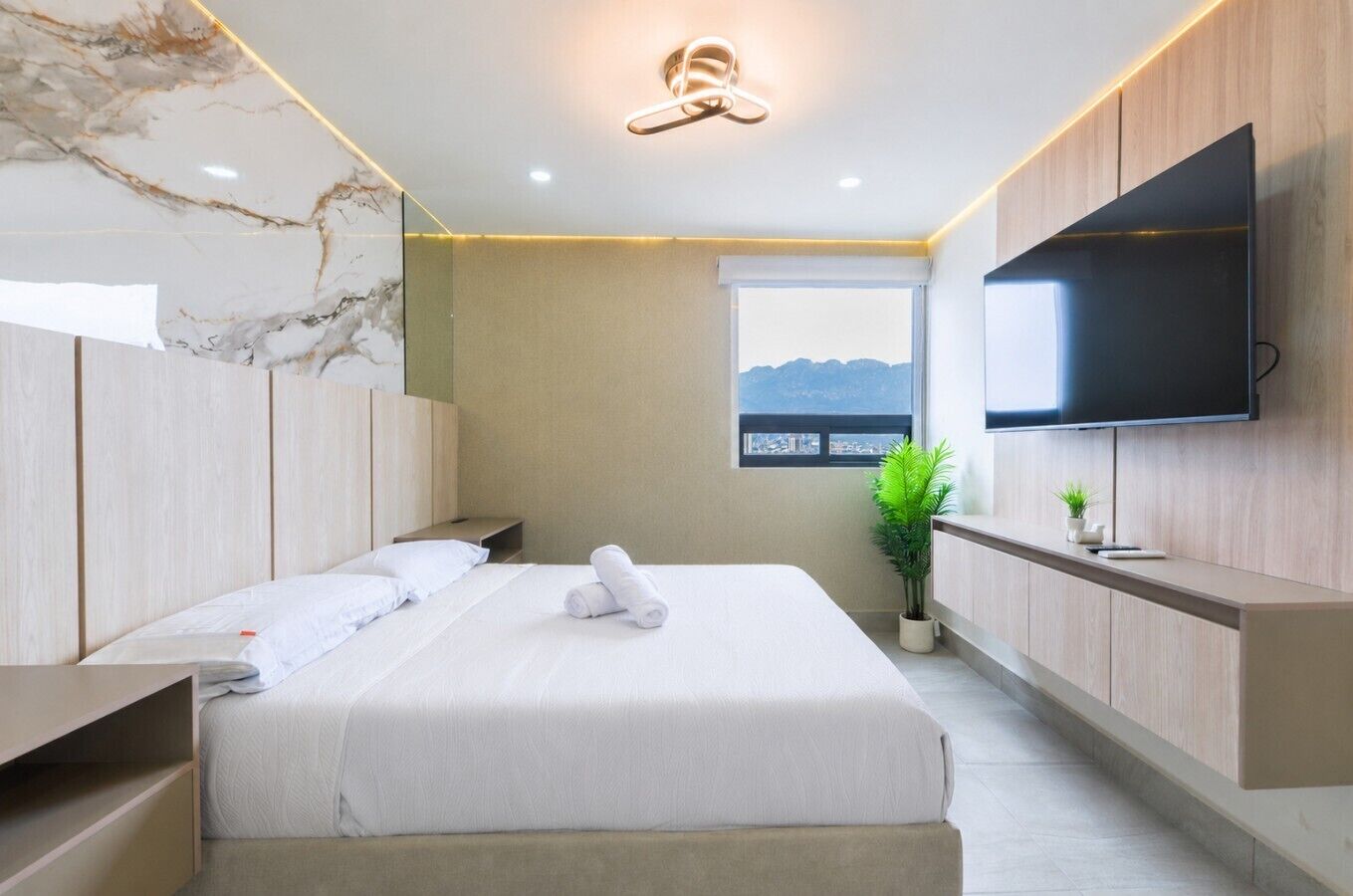 A modern hotel room with a bed, a flat-screen TV, a window with a view, and a plant.