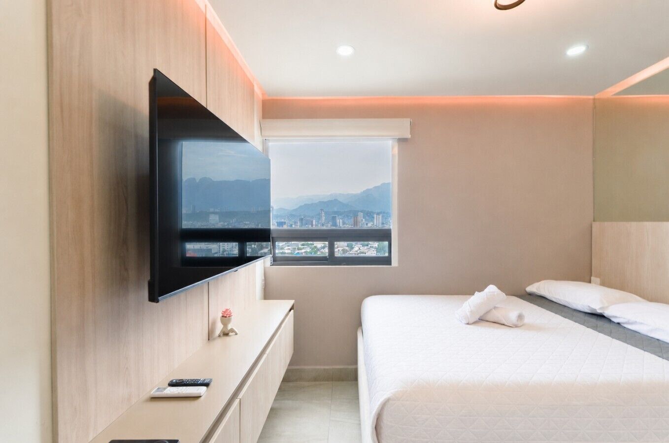 A modern hotel room with a bed, a flat-screen TV mounted on the wall, a small desk with a remote control, and a window with a cityscape view.