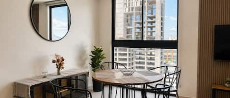 Un salon moderne avec une table à manger ronde, des chaises noires, une grande fenêtre donnant sur des immeubles, un miroir rond et un téléviseur à écran plat.