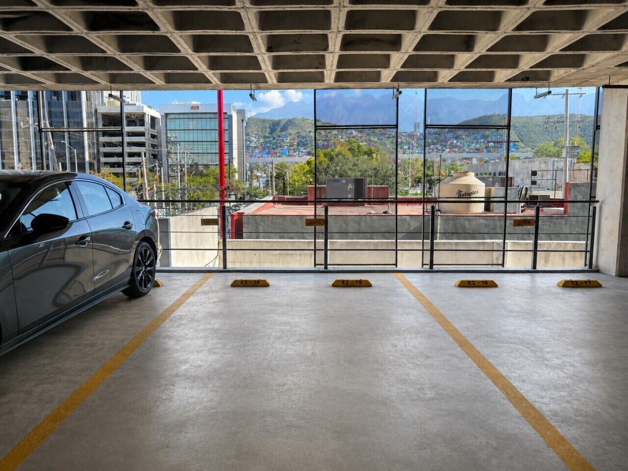 Eine Parkgarage mit einem Auto in einem markierten Stellplatz, Betonwänden und Blick auf eine Berglandschaft durch große Fenster.