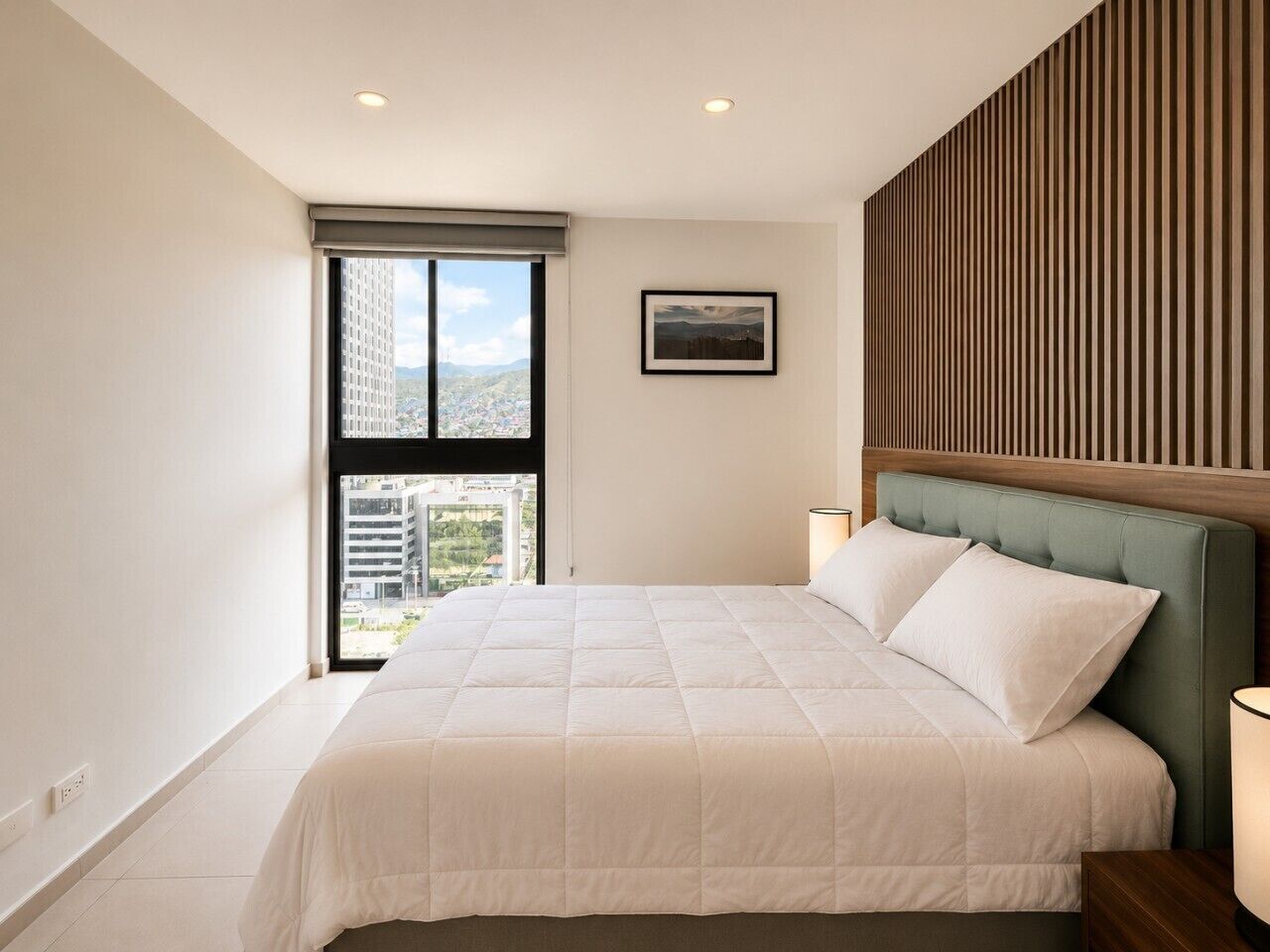 A bedroom with a large bed, a window with a view of buildings, a framed picture on the wall, and a wooden panel design on the wall.