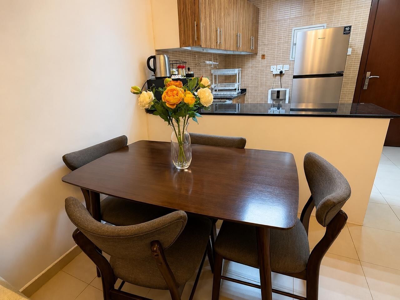 Un coin repas avec une table et quatre chaises, un vase avec des fleurs et, au fond, une cuisine.