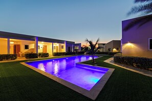 Una piscina iluminada con luz azul, rodeada de un césped cuidado y un edificio con ventanas iluminadas.