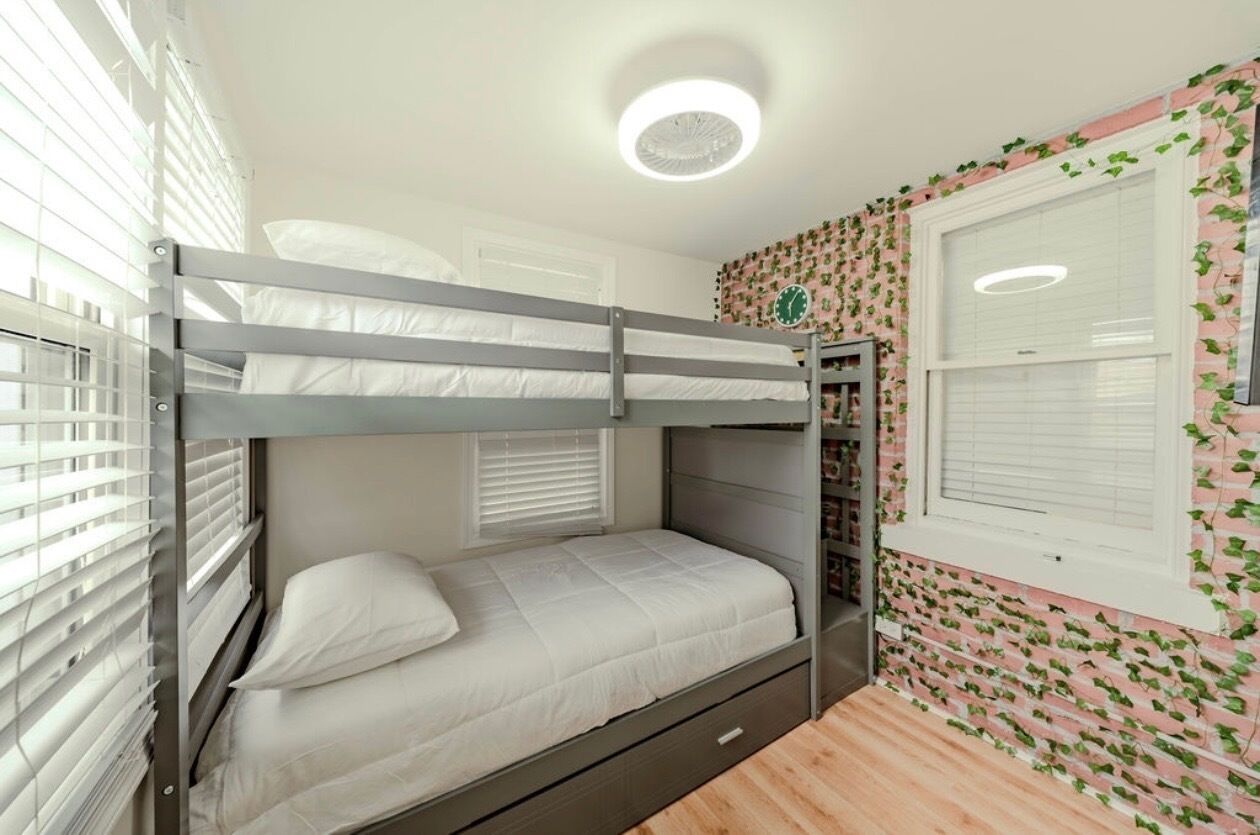 Une chambre avec un lit superposé, un luminaire au plafond, une fenêtre avec des stores et un mur orné d’un motif végétal.