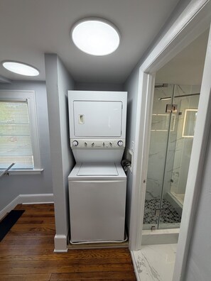 Une petite buanderie équipée d’un lave-linge et d’un sèche-linge à chargement frontal, d’une cabine de douche en verre et d’une fenêtre avec des stores.