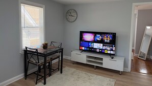 A living room with a TV, a dining table with chairs, a clock on the wall, and a window with blinds. - Modern 2BR Near Beach+Private Parking (West Haven)