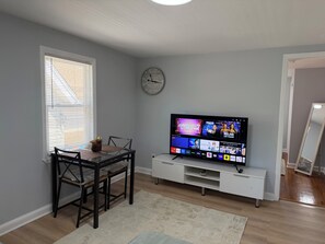 A living room with a TV, a dining table with chairs, a clock on the wall, and a window with blinds.