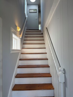 Un escalier avec des marches peintes en blanc et des contremarches en bois, menant à un palier où se trouve un luminaire et un panneau encadré.