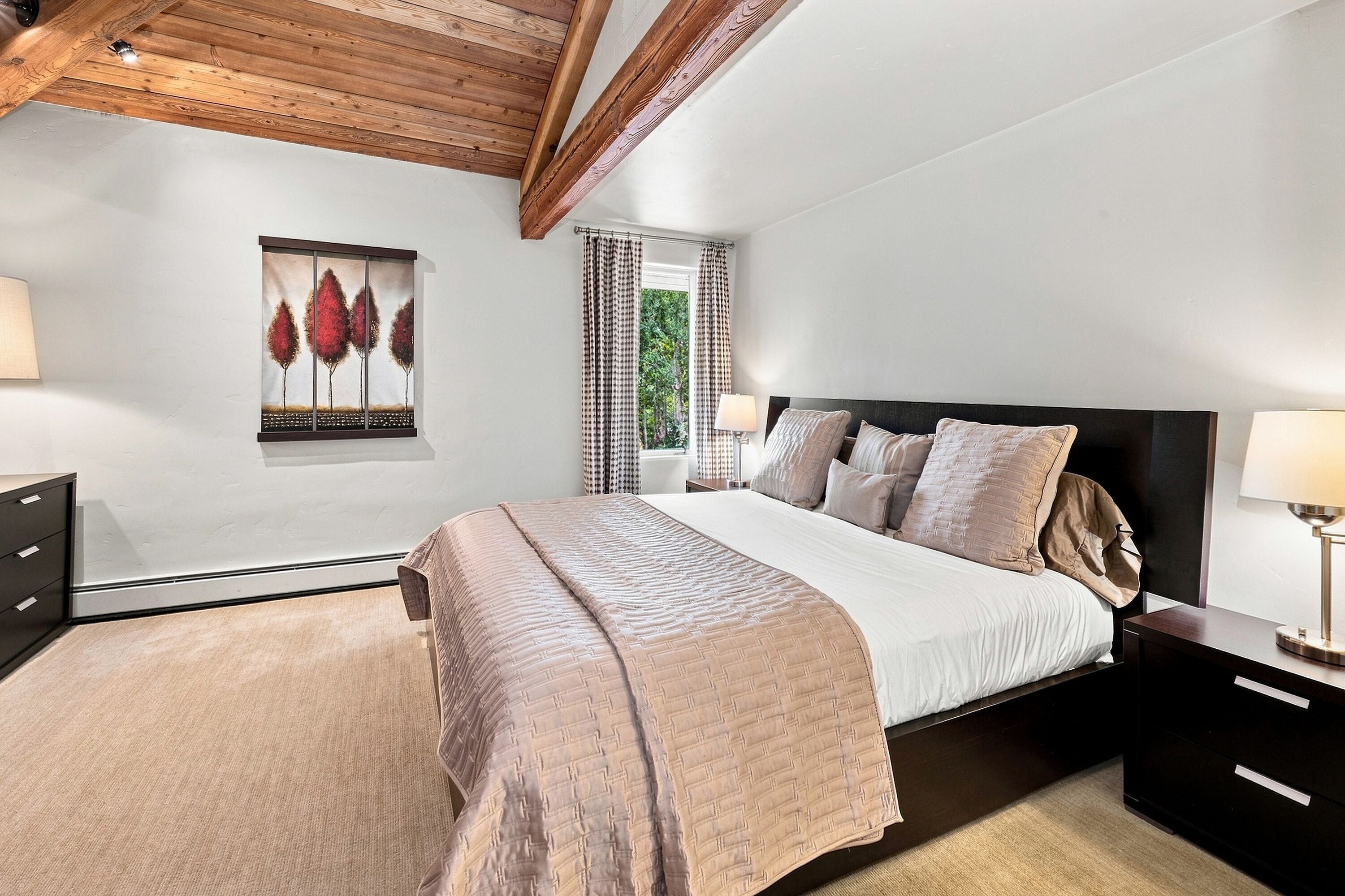 A bedroom with a bed, bedside tables, a lamp, a window with curtains, and a painting on the wall.