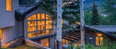 A modern house with a large glass window, surrounded by trees and a mountain in the background.