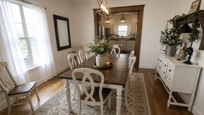 A dining area with a wooden table, white chairs, a rug, and a kitchen in the background.