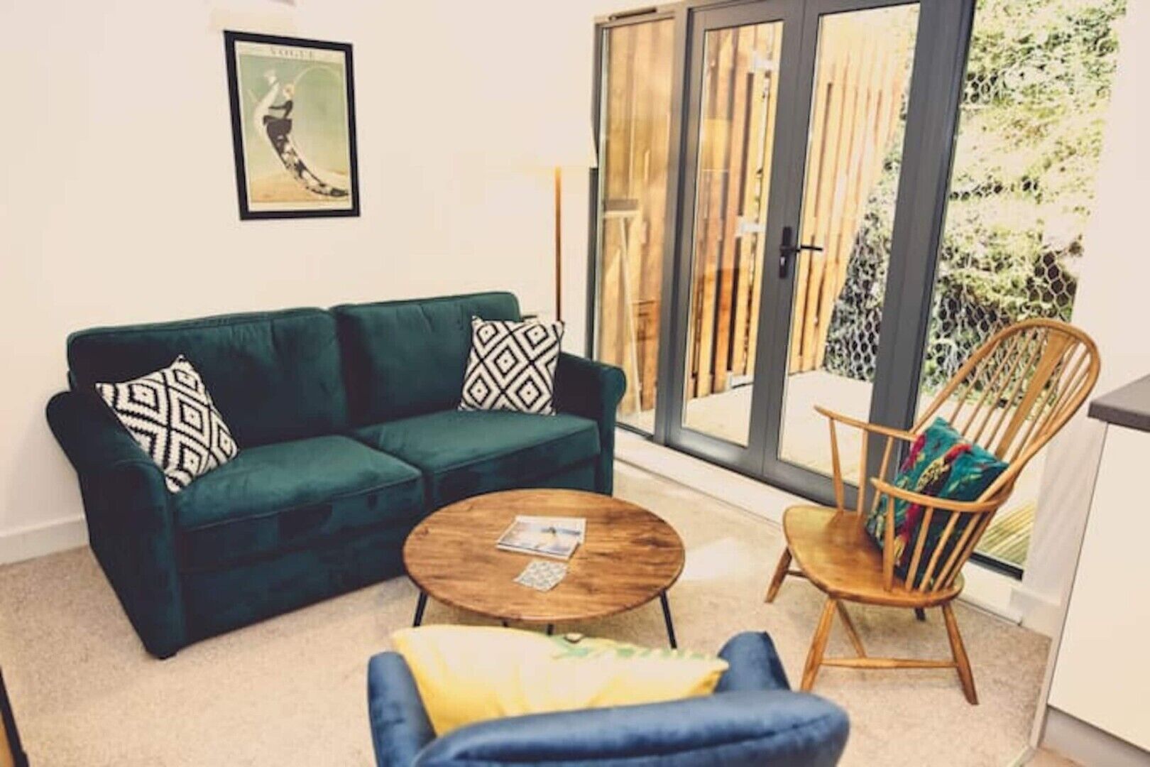 A living room with a green sofa, a round wooden coffee table, a blue armchair, and a wooden chair with colorful cushions.